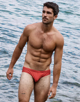 A shirtless man standing in shallow water wearing rust orange bikini swimwear.