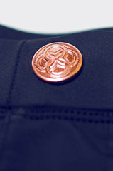 A copper-toned circular button with an intricate interlocking design on dark navy fabric of bondi short