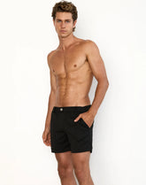 A shirtless man modeling black Trinity shorts with one hand in his pocket against a white background.