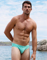 A shirtless man wearing cool mint 2EROS V10 core bikini swimwear, posing outdoors near the water.