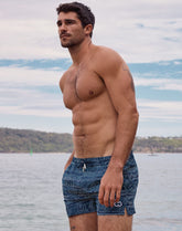 Shirtless man posing by the water, wearing blue patterned swim shorts with a drawstring waist.