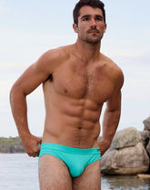 A muscular man standing near a rocky shore, wearing the 2EROS V10 Core Bikini Swimwear in neon mint.