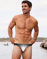 A shirtless man wearing 2EROS V20 black and white comic print swim brief by a body of water.