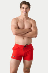 Shirtless smiling male model wearing bright red AWRY Menswear gym shorts with black drawstrings and a black logo tag.