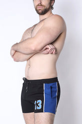 A shirtless man wearing black CellBlock 13 shorts with a blue side panel, white piping, and a light blue number 13.
