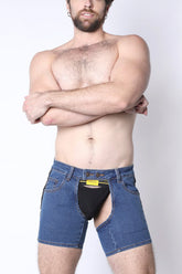A shirtless man wearing indigo denim chap shorts with an open front, revealing a black jockstrap.