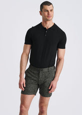 A male model stands wearing black POSTMARC chalk stripe jacquard stretch knit chino shorts and a black polo shirt.