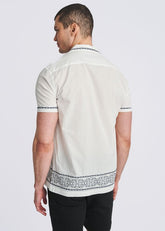 Man standing in profile wearing a white short-sleeve button-down shirt with blue medallion embroidery along the collar, cuffs, and hem.