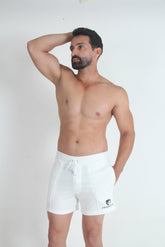 A shirtless bearded man posing against a white wall, wearing white Tulio summer fleece shorts with a drawstring.