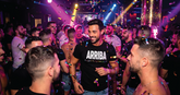 A man in a black t-shirt with ARRIBA in white text stands in a crowded, confetti-filled nightclub.