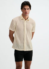 A man poses in an off-white open knit short-sleeve button-down shirt with a rib collar, paired with black shorts.