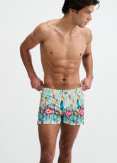 A male model wearing ST33LE teal swim shorts with a multicolored abstract floral drip pattern on a white background.