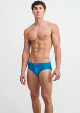A male model stands wearing ST33LE textured cobalt swim briefs featuring a gold front buckle against a white background.