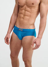 Muscular man wearing cobalt textured swim briefs with a gold front buckle against a white background.