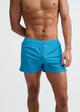 A shirtless man wearing bright blue pinstripe swim shorts against a plain white background.