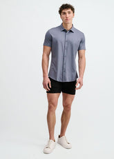 A male model wears a gravel-colored short-sleeve stretch knit button-down shirt with black shorts and white sneakers.