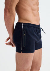A shirtless man in profile wears navy gym shorts with white piping, a drawstring waist, and a zippered pocket.