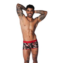 Camouflage print trunk with red trim and branded elastic waistband, worn by a tattooed model on a black background.