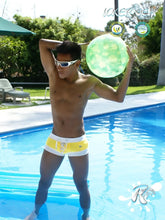 A man stands in a pool wearing icker sea legs mango swim trunks holding a translucent green inflatable ball
