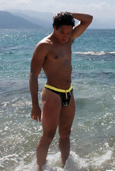 A man stands in ocean waves wearing icker sea letters thong swimwear in black and mango with a yellow waistband