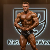 A muscular male bodybuilder wearing black shiny MetroMaleWear crocodile swim briefs with silver number 3 badge on waistband