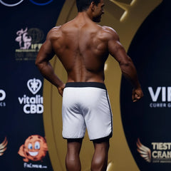 A muscular man stands on stage wearing white MetroMaleWear MetroMuscleWear Avalanche Physique board shorts