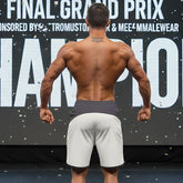 A muscular man stands on stage wearing gray metro musclwear cordoba physique board shorts with dark gray waistband
