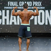 A muscular man wearing navy blue MetroMaleWear MetroMuscleWear Marine Physique board shorts with light green waistband and pocket