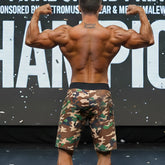 A muscular man wearing metroMalewear physique board shorts in green brown and beige camouflage pattern