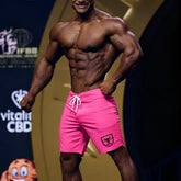 Bright pink MetroMaleWear physique board short with white drawstring and black shield logo on lower left leg