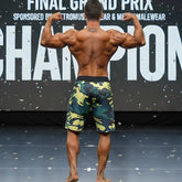 A muscular man stands on stage wearing MetroMaleWear physique board shorts in yellow green camouflage pattern