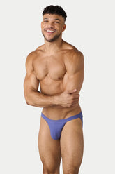 A smiling man models the AWRY Menswear lavender Oaisis swim tanga against a plain light background.