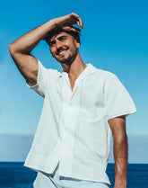 White santorini linen shirt with short sleeves, light blue embroidery along seams, and logo on left chest