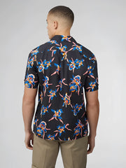 A man wearing a dark navy short-sleeved button-up shirt with vibrant blue and pink floral patterns signature botanical print