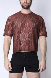 A bearded man wearing a rust-colored lace cropped tee and black shorts against a plain background.