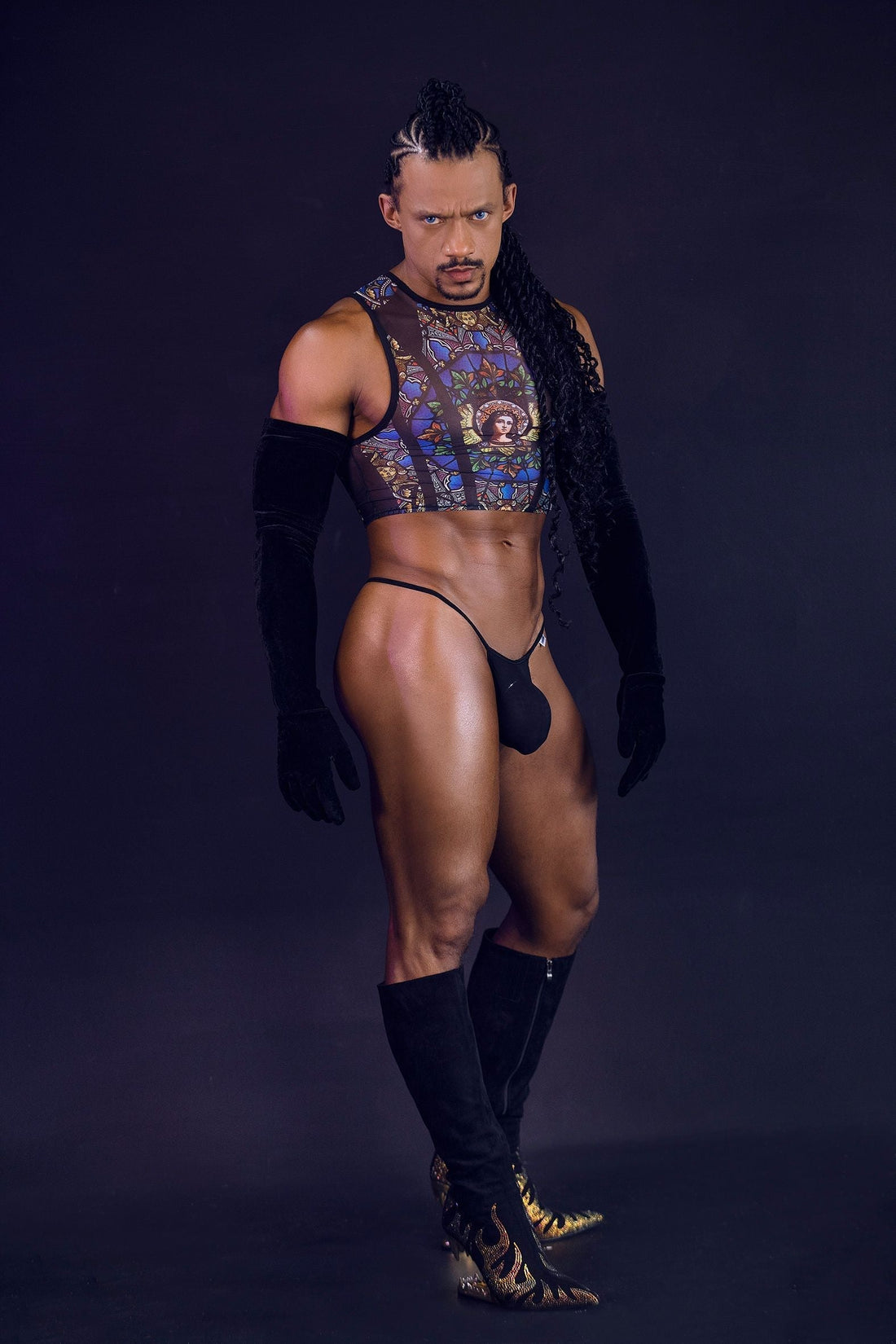 Muscular man in a bold, colorful crop top, black thong, long gloves, and ornate boots.