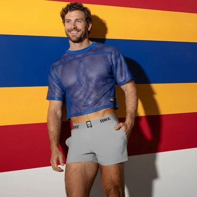 BIKE athletic wear lifestyle shot of male model in blue mesh crop top tee and grey gym shorts against striped wall