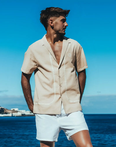 Man wears a beige short-sleeve shirt and white shorts near blue water.