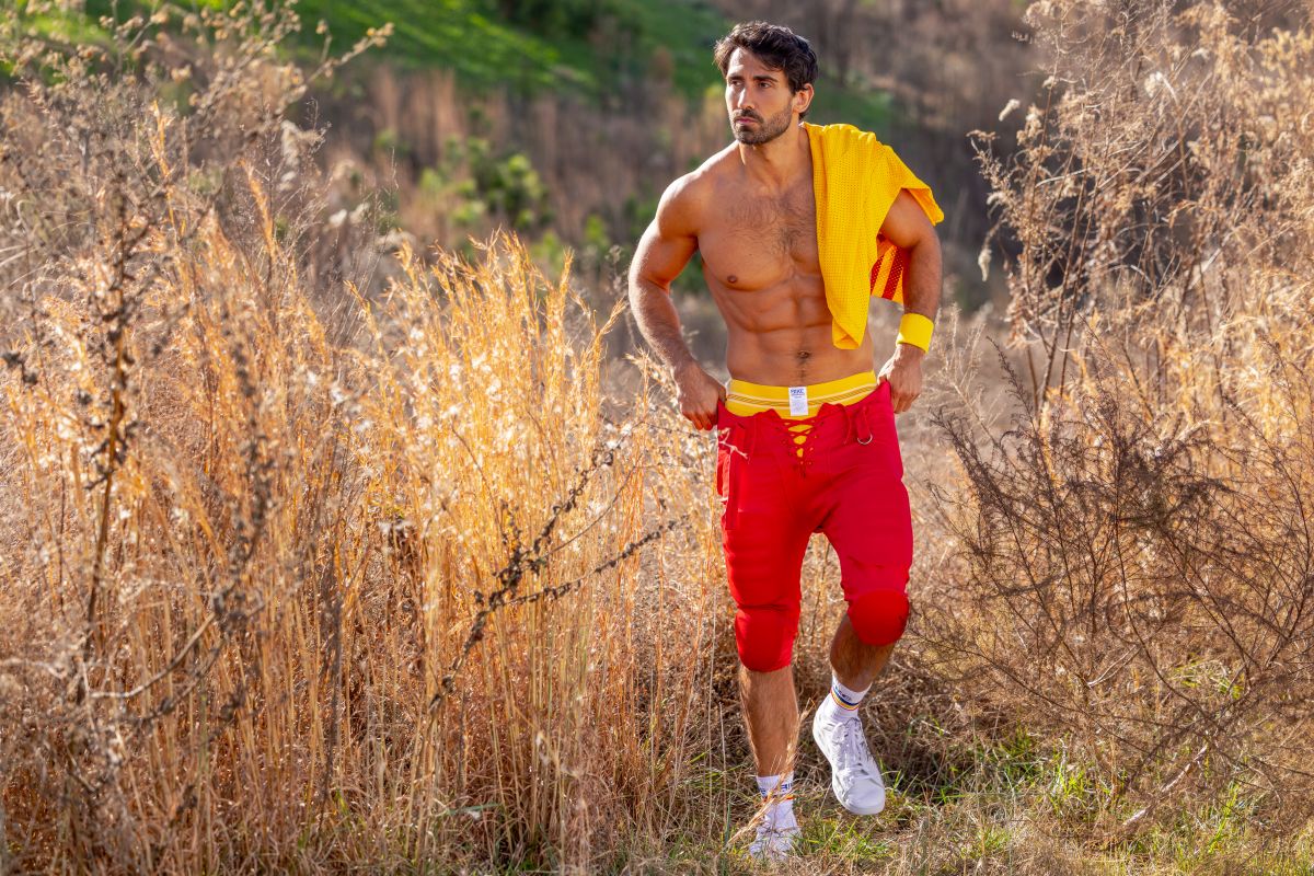A muscular man wearing a yellow bike athletic BAS306 original number 10 jockstrap with red shorts and white sneakers