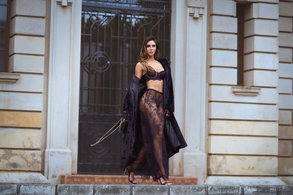 A black three-piece lingerie set featuring a lace bra, matching thong, and a sheer lace maxi skirt, styled with a coat.