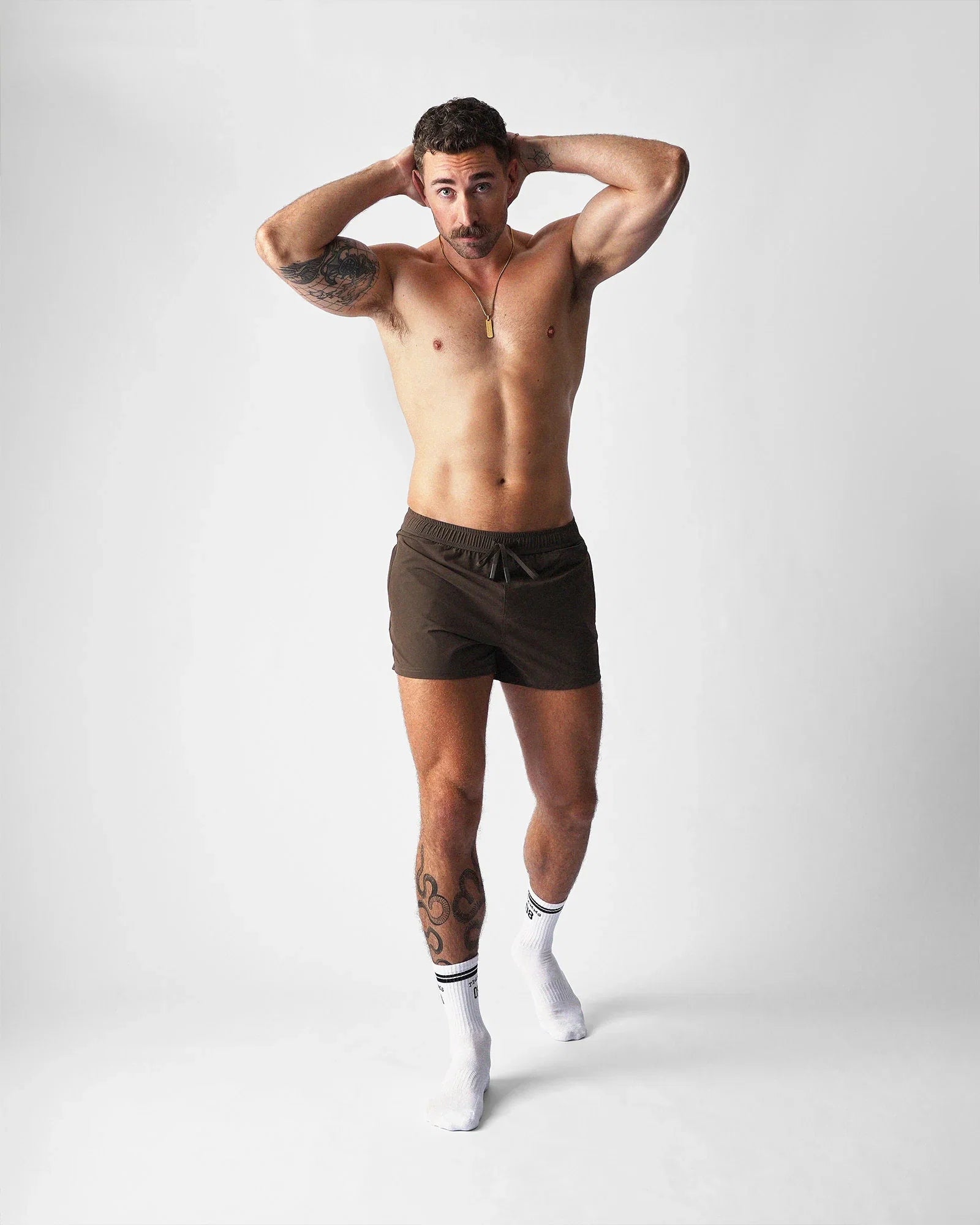 A muscular man wears teamm8 brown gym shorts, white socks, and hands behind his head on a light background.