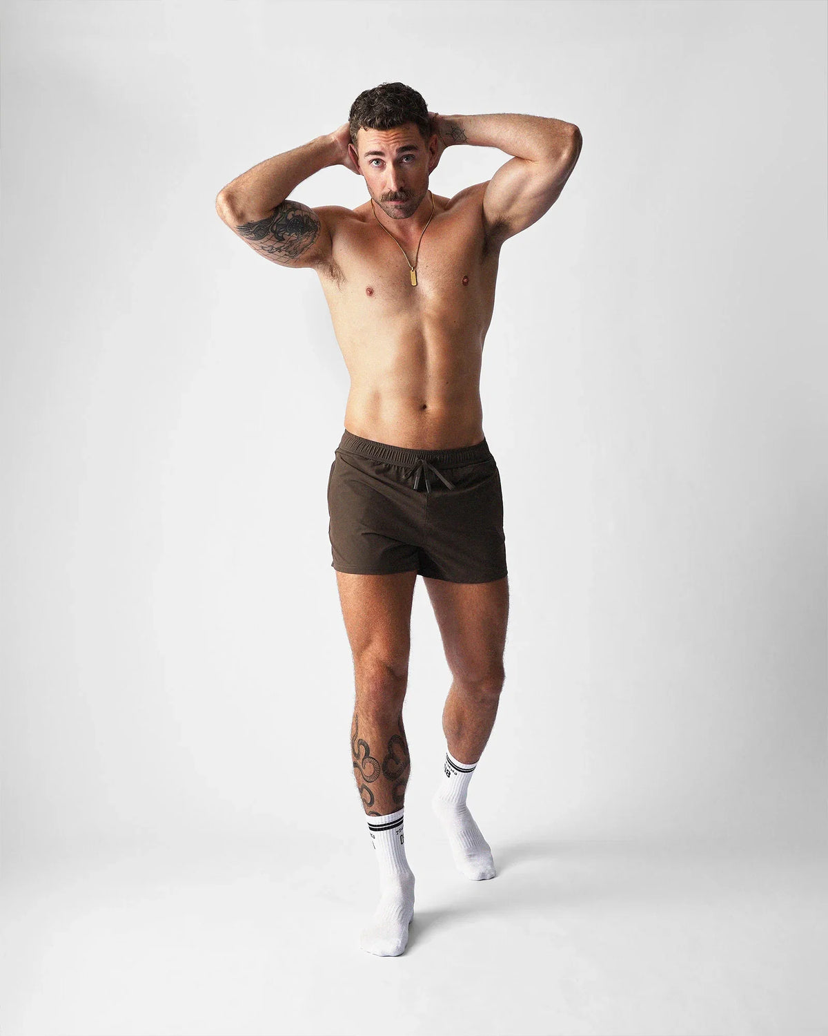 A muscular man wears teamm8 brown gym shorts, white socks, and hands behind his head on a light background.