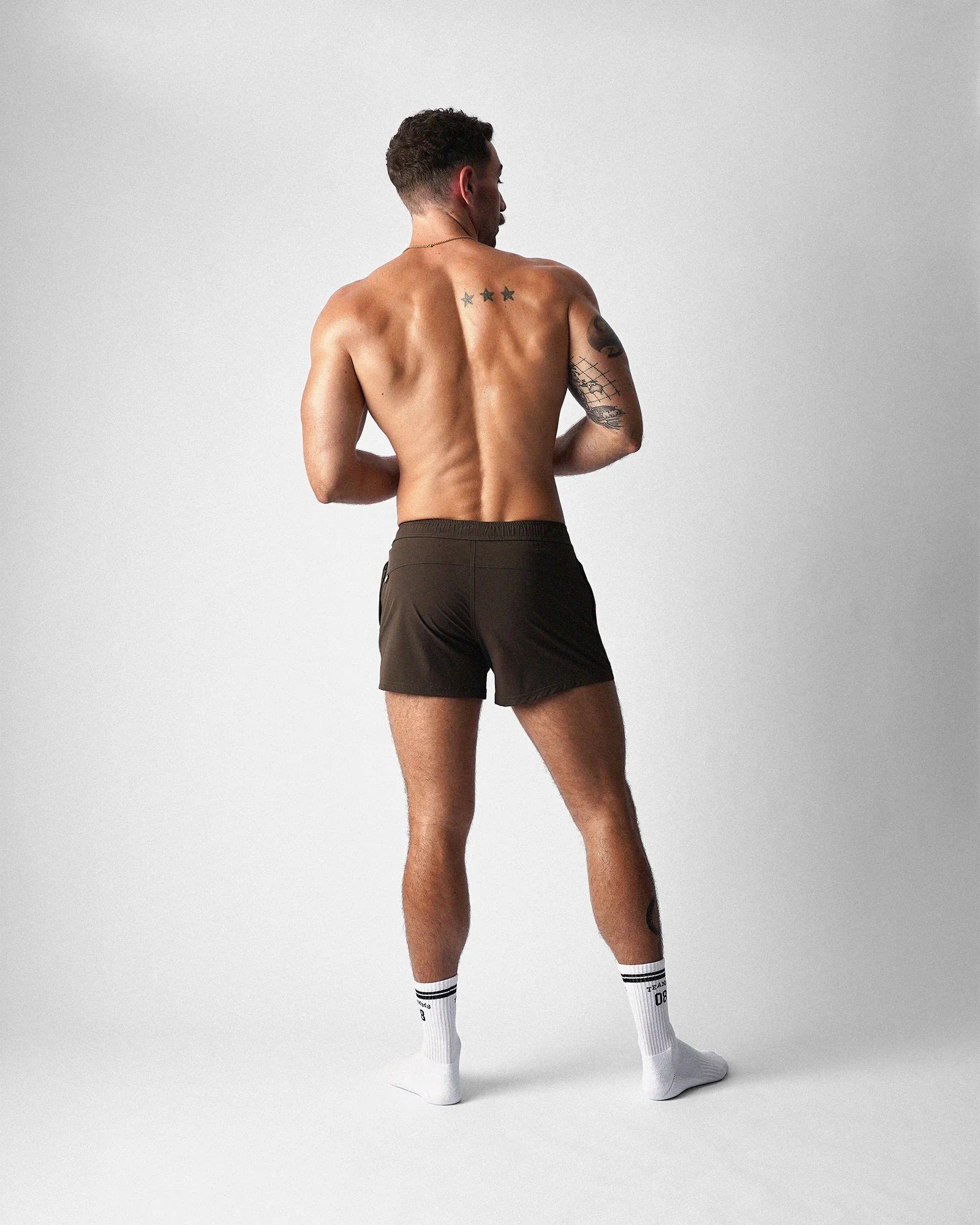 Rear view of a muscular male model wearing brown gym shorts and white socks against a plain white background.