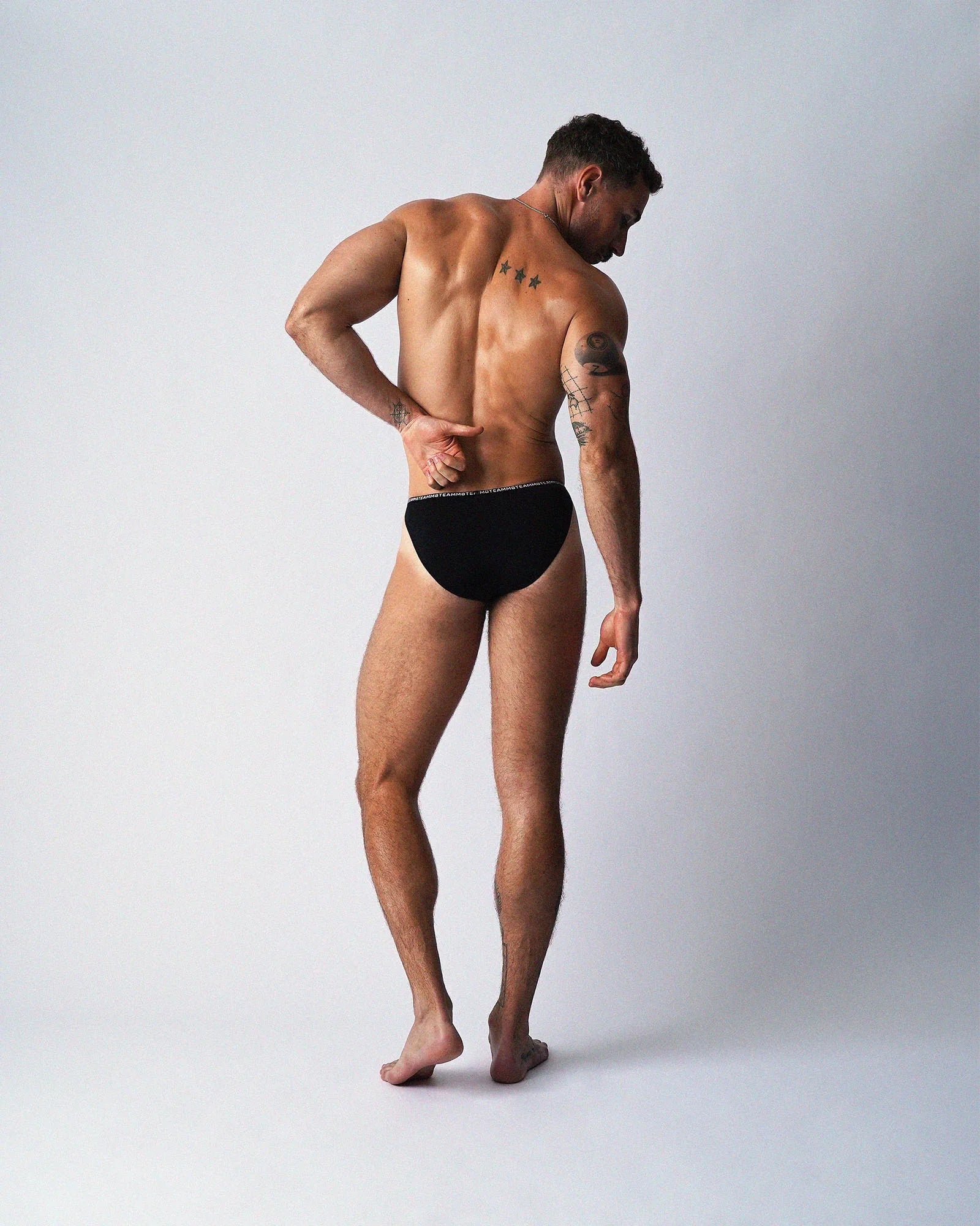 A muscular man wearing black teamm8 Glide Micro Brief underwear, viewed from the back against a plain background.