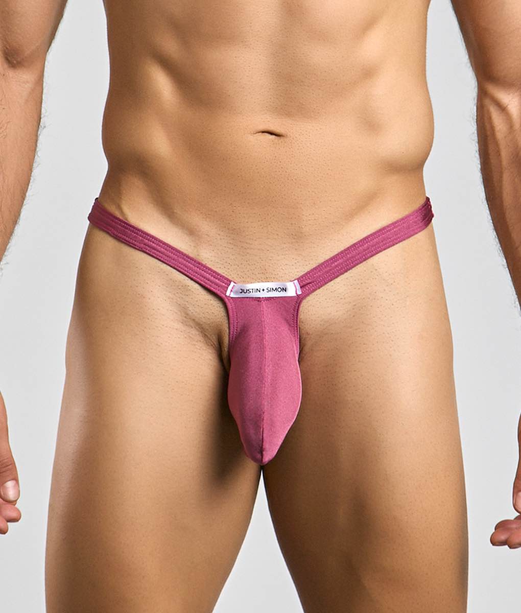 A magenta form-fitting thong with a central pouch and a white label featuring the justin + simon brand name