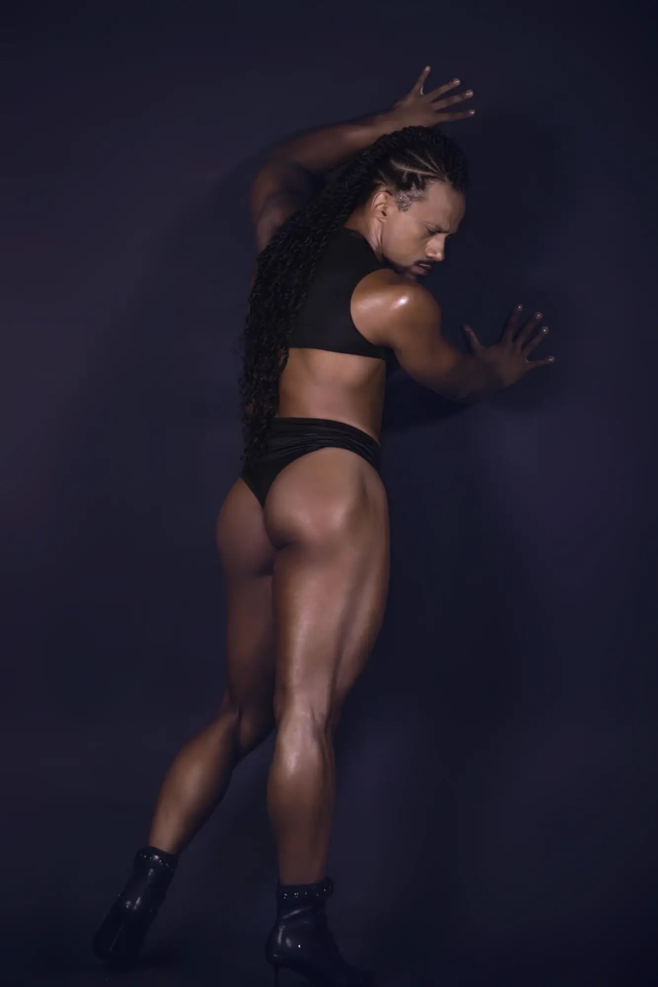 A muscular individual wearing the black CandyMan 99783 one shoulder bodysuit paired with glossy black heeled boots