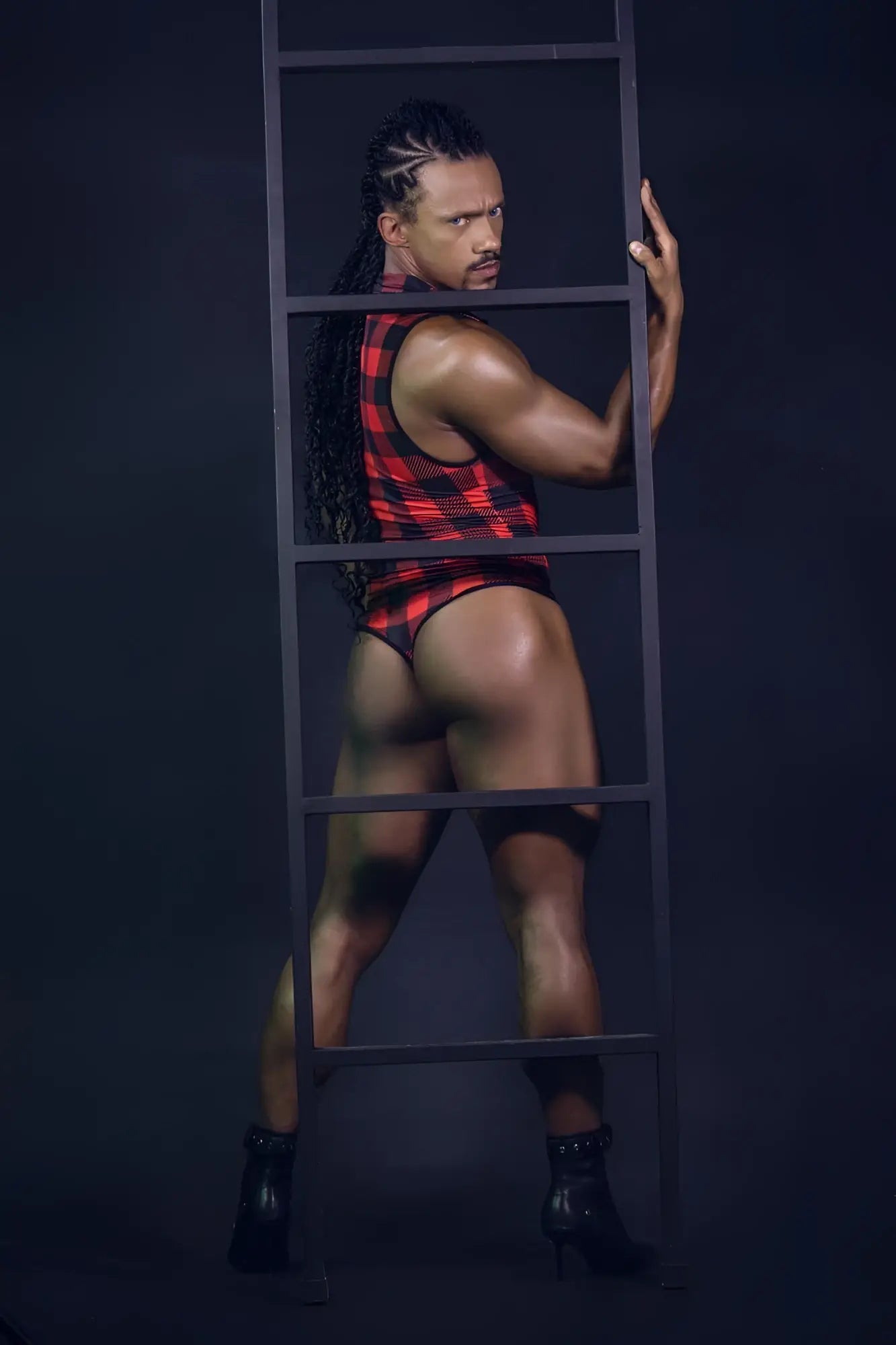 A muscular man with long braided hair wears a red and black plaid sleeveless lumberjack bodysuit and briefs standing behind a ladder