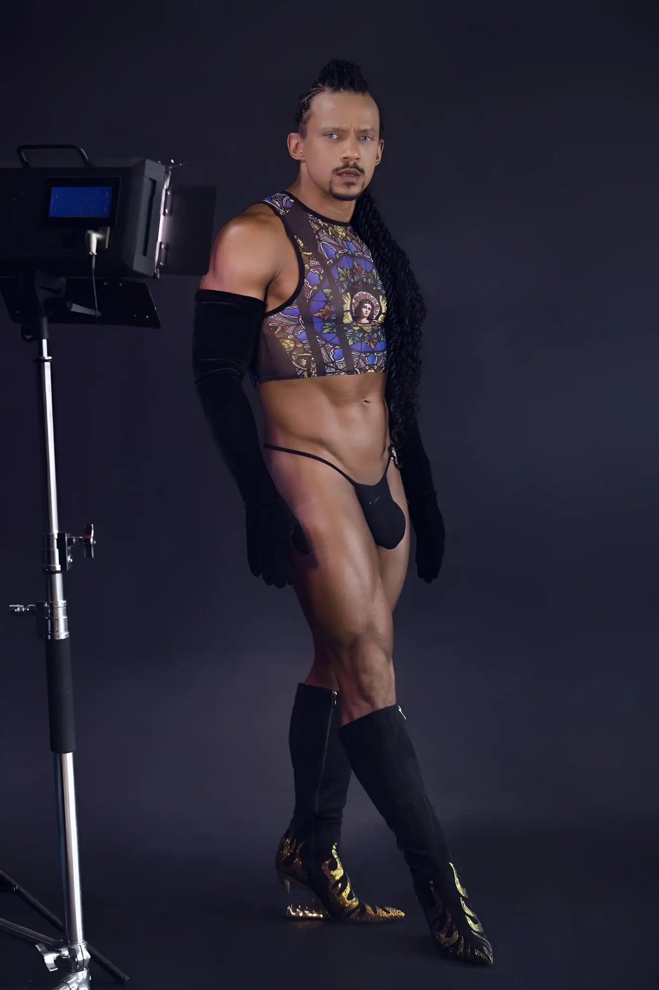 A man wearing a black candyMan 99805 stained glass tank top with gold patterned mesh and matching thigh-high boots