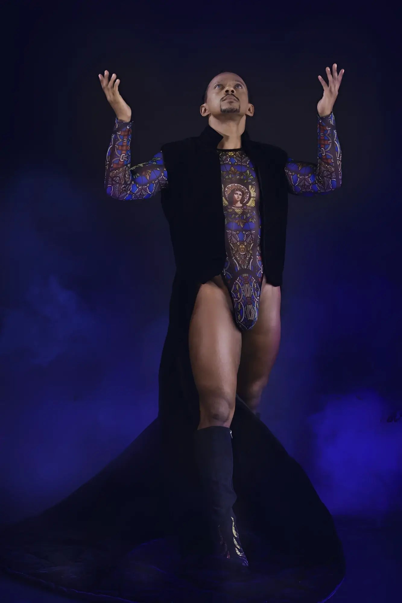 A man wearing a black vest over a candyMan 99814 stained glass bodysuit with blue and gold patterns