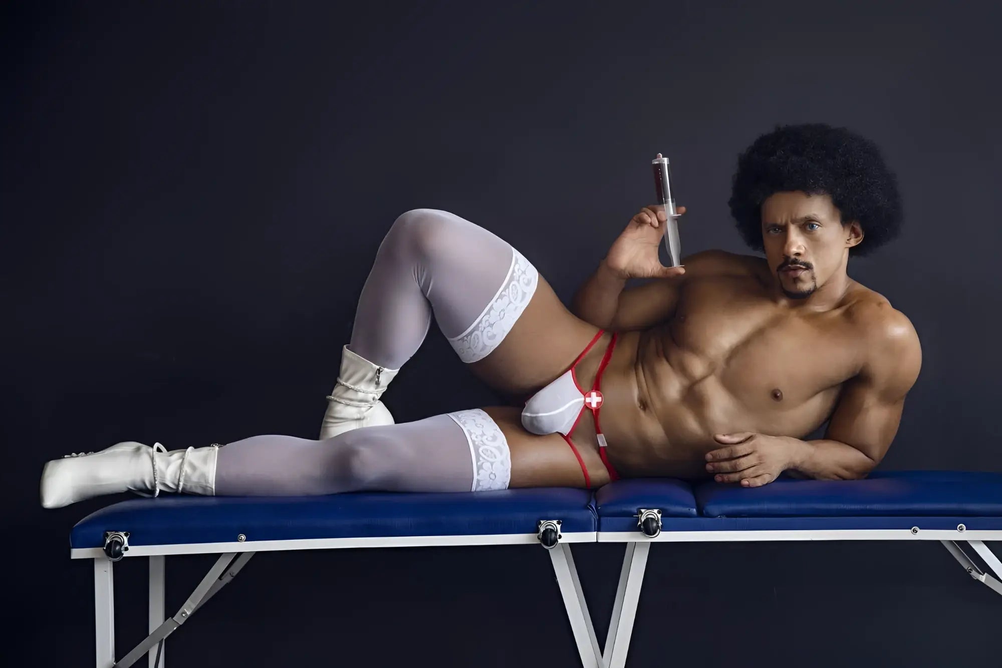 A muscular man with afro lies on blue table wearing CandyMan 99833 murse thongs red-white and white lace thigh-high stockings